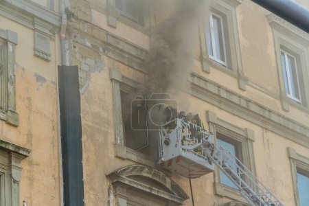 Photo for Naples, Italy - February 15th 2023 - Domestic apartment fire with smoke billowing from window. Garibaldi Square, Naples, Italy. Fire engine basket or cage with two firemen, , one preparing to enter. - Royalty Free Image