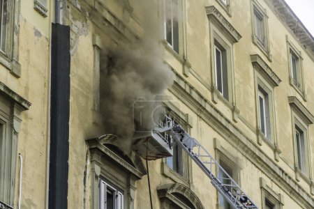 Photo for Naples, Italy - February 15th 2023 - Domestic apartment fire with smoke billowing from window. Garibaldi Square, Naples, Italy. Fire engine basket or cage with two firemen in breathing apparatus, landscape. - Royalty Free Image