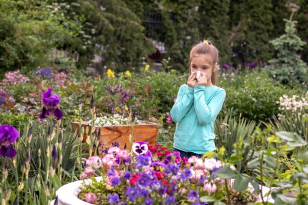 Allergien. Ein kleines Mädchen pustet seine Nase in ein Taschentuch, schockiert von einer allergischen Reaktion auf blühende Blumen in der Nähe eines Blumenbeetes. Allergie-Konzept. Kopierraum.