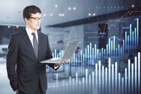 Attractive thoughtful young european businessman using laptop with abstract glowing and growing business graph hologram on blurry office interior background. Planning and strategy, stock market, business growth, progress and financial success concept