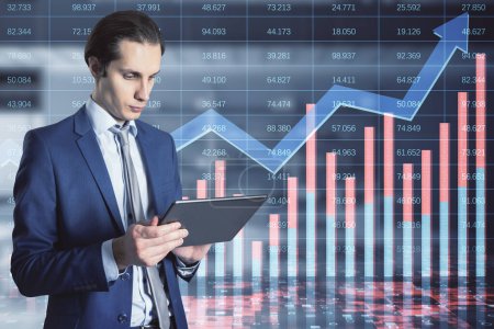 Attractive young european businessman using tablet with creative growing financial forex chart with upward arrow on blurry office interior background. Market, stock and trading concept. Double exposure