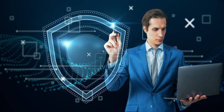 Attractive young caucasian businessman in suit and tie holding laptop with creative glowing shield hologram on blue background with various blurry icons. Cyber security, data protection concept. Modern wireframe design