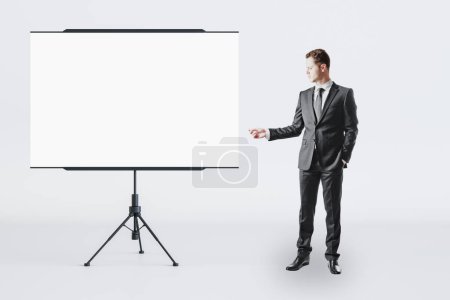 Business education and teamwork concept with businessman front view near blank white flip chart with place for your logo or text in abstract room on light wall background, mockup