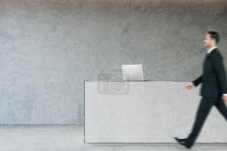 Side view of young business man walking in modern concrete office reception interior with desk. Lobby concept. Worker and CEO concept