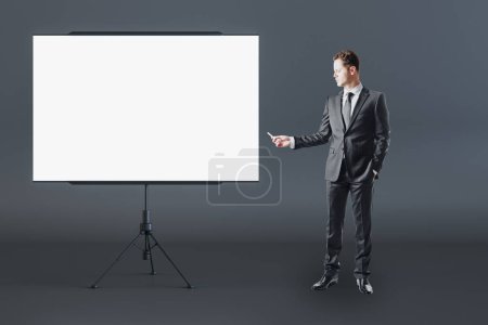 Business education and teamwork concept with man in black suit front view near blank white flip chart with place for your logo or text in abstract room on dark wall background, mockup