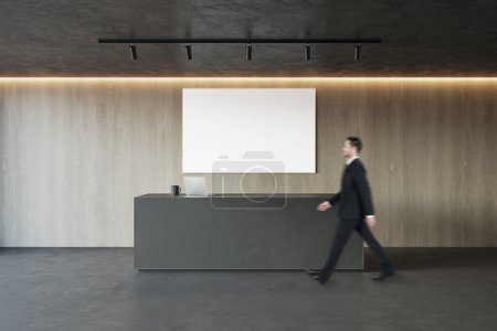 Side view of young businessman walking in modern concrete and wooden office reception interior with empty white mock up banner and desk. Lobby concept. Worker and CEO concept
