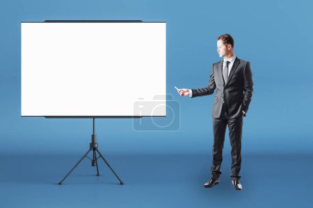 Business Education-Konzept mit Geschäftsmann-Frontansicht mit Kreide in der rechten Hand in der Nähe eines leeren weißen Flipcharts mit Platz für Ihr Logo oder Text im abstrakten Raum auf blauem Wandhintergrund, Attrappe