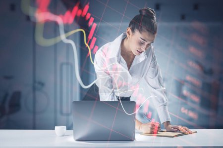 Photo for Attractive blonde young european businesswoman using laptop at desktop with abstract falling red forex chart on blurry office interior background. Economic decline and loss concept - Royalty Free Image