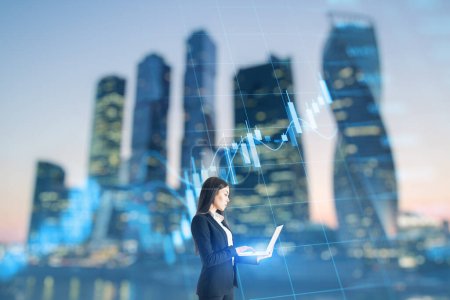 Trade, finance and market concept. Attractive young caucasian businesswoman using laptop on blurry city background with forex chart. Double exposure