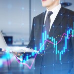 Young european businessman holding laptop with glowing candlestick forex chart hologram on blurry office interior background. Trade and market concept. Double exposure