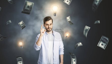 Erfolgreiche Geschäftsidee, Geldverdienen und Kreativität Konzept mit gutaussehendem Mann mit erhobenem Zeigefinger auf nebligem Hintergrund mit Glühbirnen und Dollarnoten