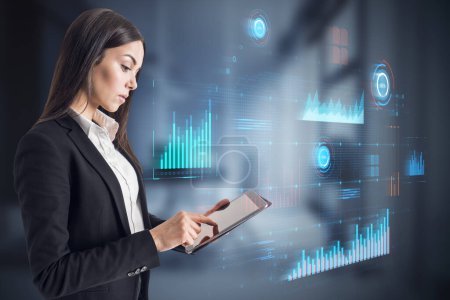 Photo for Attractive blonde businesswoman using tablet with glowing digital business interface with charts on blurry office interior background. Infographic and big data concept. Double exposure - Royalty Free Image