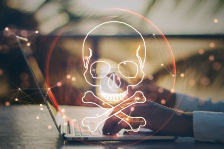 Close up of male hand using laptop and cellphone at desktop with digital red skull on blurry background. Ransomeware, virus and pirate threat concept. Double exposure