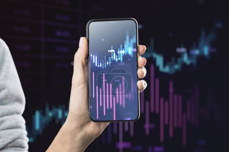 Close up of female hand holding mobile phone with glowing candlestick forex chart on blurry background. Investment, profit and financial growth concept. Double exposure