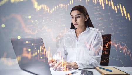 Attrayant jeune femme d'affaires en utilisant un ordinateur portable au bureau avec hologramme de chandelier forex rouge vers le bas sur fond flou. Crise financière, stock et concept de récession. Double exposition