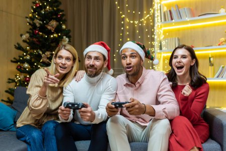 New Years party group of four diverse friends having fun relaxing and celebrating on Christmas holidays, guests sitting on sofa men and women playing video games on joystick consoles.
