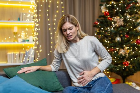 Eine kranke Frau sitzt allein zu Hause, hat zu Weihnachten starke Bauchschmerzen in der Nähe des Weihnachtsbaums an Silvester.