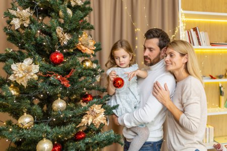 Téléchargez les photos : Une jeune famille heureuse décore un sapin de Noël pour les vacances de Noël. Papa tient sa fille dans ses bras, la fille pend un jouet, maman étreint papa. - en image libre de droit