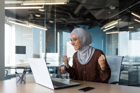 Successful businesswoman boss celebrating victory and successful triumph, muslim in hijab looking at laptop screen and holding hand up gesture of success and achievement, woman working inside