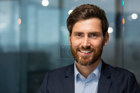 Photo for Closeup photo headshot of a mature businessman with a beard looking at the camera and smiling, a successful investor banker inside a modern office building at work, a man in a business suit evening - Royalty Free Image