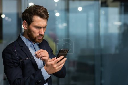 Mature businessman in office near window in evening upset reading message online notification from phone, male boss working inside office in business suit and glasses with beard, upset investor.