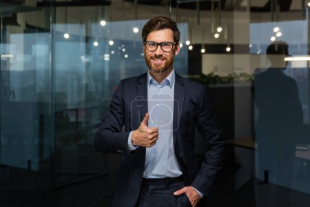 Photo for Portrait of successful mature businessman boss, manager in business suit glasses and beard looking at camera and smiling standing near window, showing thumbs up, sign of success and achieving goals. - Royalty Free Image
