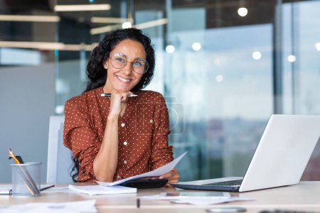 Photo for Portrait of happy and successful hispanic woman, businesswoman smiling and looking at camera holding contracts and invoices, working inside office with laptop on paper work. - Royalty Free Image