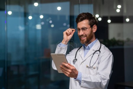 Foto de Un joven médico, interno, interno está de pie en un consultorio en un hospital con una bata blanca y con un estetoscopio. Él trabaja en una tableta, sostiene sus gafas en su mano, y sonríe. - Imagen libre de derechos