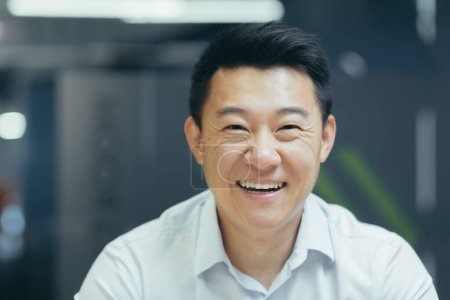 Fotografía de cerca. Retrato de un apuesto joven empresario asiático. Él está sentado en la oficina con una camisa blanca, mirando a la cámara, sonriendo.