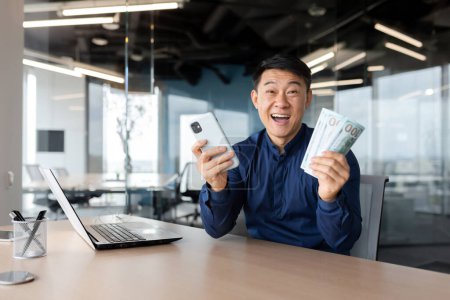 Téléchargez les photos : Portrait homme d'affaires réussi asiatique souriant contentement et regardant la caméra tenant téléphone et argent cash dollars dans les mains, célébrant la victoire, homme en chemise travaillant à l'intérieur du bureau en utilisant un ordinateur portable. - en image libre de droit