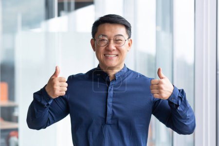 Foto de Retrato de un hombre asiático exitoso dentro de la oficina cerca de la ventana, jefe en gafas y camisa mirando a la cámara y mostrando los pulgares hacia arriba. - Imagen libre de derechos