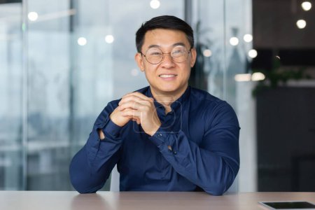Young successful Asian businessman looking at caper and smiling, video call sitting at table investor boss in shirt and glasses.