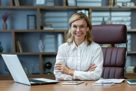 Erfolgreiche Unternehmerin im Hemd sitzt mit verschränkten Armen lächelnd am Tisch und blickt in die Kamera, Anwältin arbeitet im modernen Büro mit Laptop bei der Arbeit.