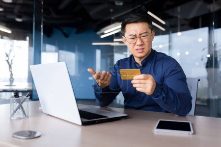 Betrügerischer Geschäftsmann, enttäuschter Mann, der im Büro sitzt, asiatischer Mann, der versucht, Banküberweisung zu tätigen und im Online-Shop zu kaufen, zu wenig Geld mit Kreditkarte.