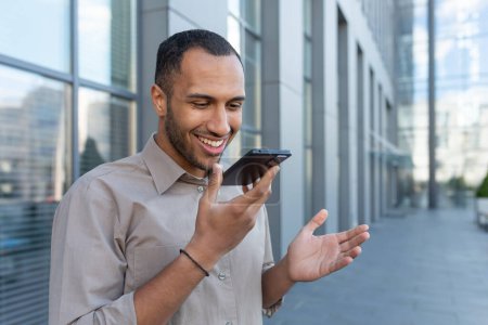 Téléchargez les photos : Un homme d'affaires hispanique souriant enregistrant un message vocal, un homme heureux devant un immeuble de bureaux lisant des commandes sur smartphone pour la recherche d'IA. - en image libre de droit