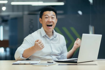 Foto de Un joven empresario asiático juega juegos en línea en el trabajo en la oficina con un ordenador portátil, realiza apuestas. Él está feliz de ganar, grita con felicidad, mira a la cámara. - Imagen libre de derechos