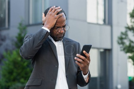 Téléchargez les photos : Mature patron afro-américain senior lisant de mauvaises nouvelles au téléphone, homme d'affaires en costume d'affaires à l'extérieur de l'immeuble de bureaux en utilisant smartphone bouleversé et déçu par la notification. - en image libre de droit
