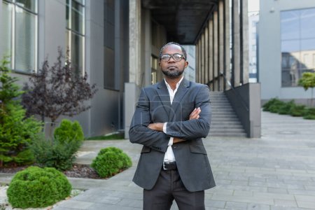 Foto de Hombre afroamericano serio y confiado en traje de negocios mirando a la cámara, hombre de negocios con brazos cruzados fuera del edificio de oficinas. - Imagen libre de derechos