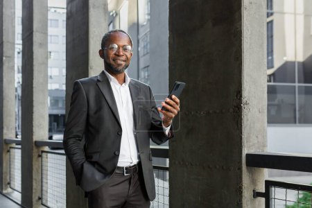Foto de Portrait of African American mature businessman, senior man outside office building holding phone in hands smiling and looking at camera. - Imagen libre de derechos