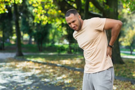 Photo for Tired man after jogging in park, hispanic man has severe pain in back and muscles after fitness, massaging his side with hand near trees. - Royalty Free Image