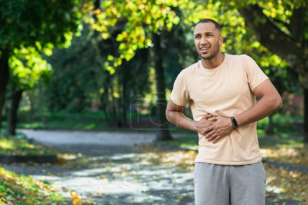 Foto de Latin american runner sportsman has severe stomach pain, man holding hand on side of stomach after doing exercise and fitness in park outside. - Imagen libre de derechos