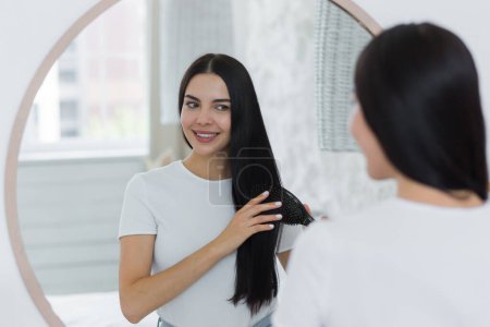 A beautiful young woman stands and looks at herself in the mirror. She combs her hair with a brush, does her hair. She smiles.