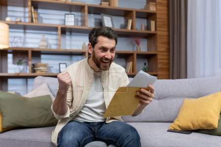 Joyful man at home holding envelope with notification message reading and smiling happy with news sitting on sofa inside living room.