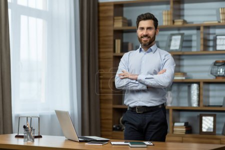 Portrait of successful businessman in office, man in shirt smiling and looking at camera, mature boss with beard with shaggy hands standing at workplace inside building.