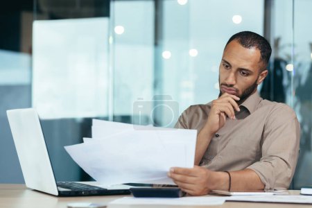 Pensive serious businessman reading financial report, hispanic businessman holding document in hands looking disappointed, working inside modern office with laptop behind paper work.