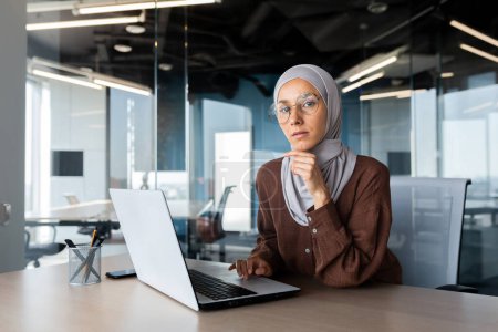 Foto de Portrait of a young woman Arab female student in hijab sitting in the office at the table and studying online at the laptop. He looks seriously at the camera. He holds his chin with his hand. - Imagen libre de derechos