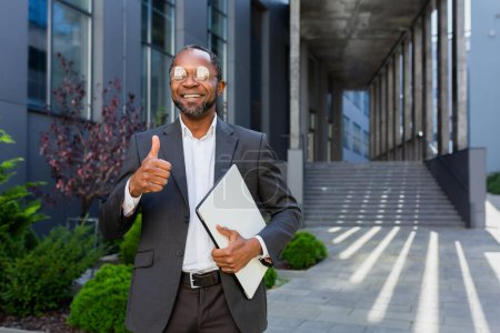 Téléchargez les photos : Portrait of successful african american man outside office building, man in suit and glasses standing and looking at camera smiling and giving affirmative thumbs up. - en image libre de droit