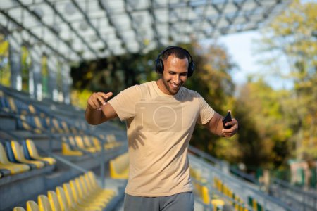 Téléchargez les photos : Young man walking in stadium after exercise and jogging in headphones, listening to audiobook music and online radio podcast, dancing joyfully, hispanic man on jogging day. - en image libre de droit