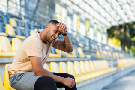 Foto de Tired and upset sportsman athlete sitting on the stadium, African American man dissatisfied with the result of training, man resting after active physical exercises and fitness. - Imagen libre de derechos