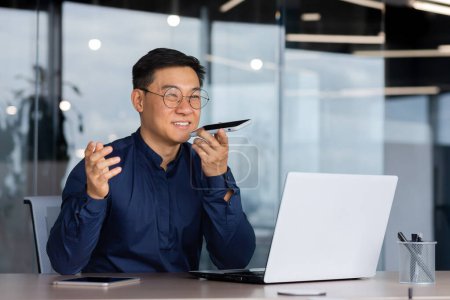 Succès asiatique homme à l'intérieur du bureau de travail avec ordinateur portable, homme d'affaires enregistrement message audio en utilisant l'application sur smartphone, l'homme en utilisant assistant d'intelligence artificielle pour aider à trouver une solution.